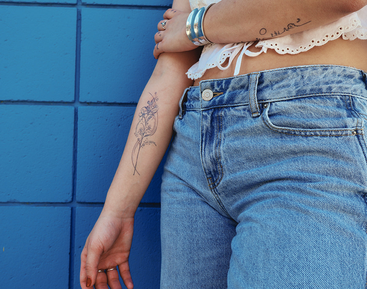 A close-up lifestyle shot of a person wearing light wash blue denim jeans and a white lace-trim top, leaning against a bright blue tiled wall. The person has temporary floral tattoos on their arms and silver bracelets on one wrist.