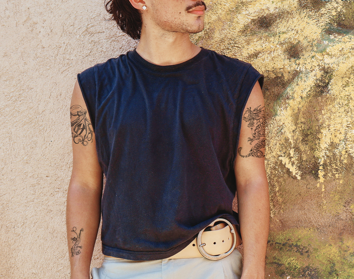 A man wearing a dark navy blue sleeveless muscle t-shirt, temporary tattoos, styled with a light beige belt and white pants, standing against a textured beige and gold stucco wall.