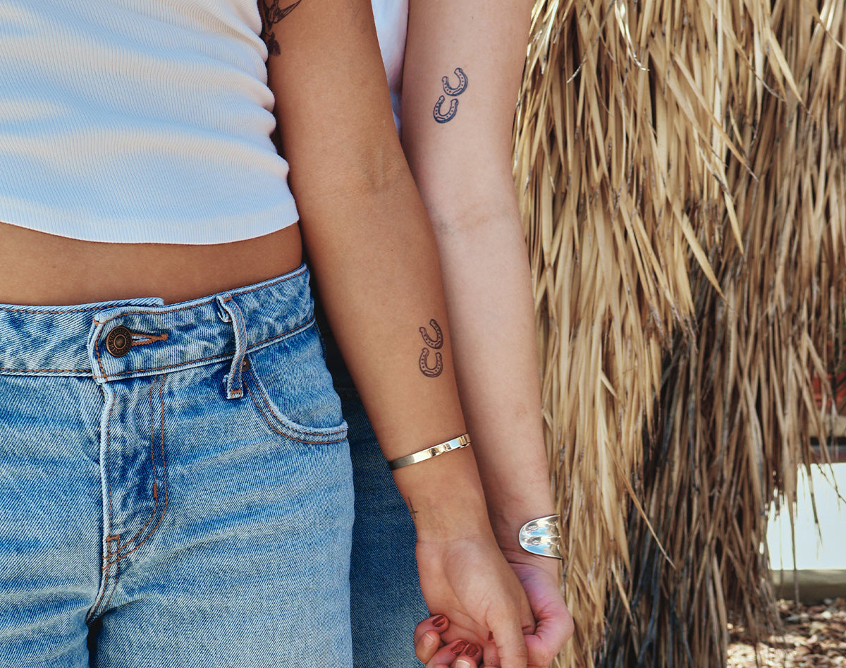 Two people standing close together outdoors, holding hands. The person on the left wears a white ribbed crop top and light blue jeans, and the person on the right wears a white sleeveless top and a silver cuff bracelet.