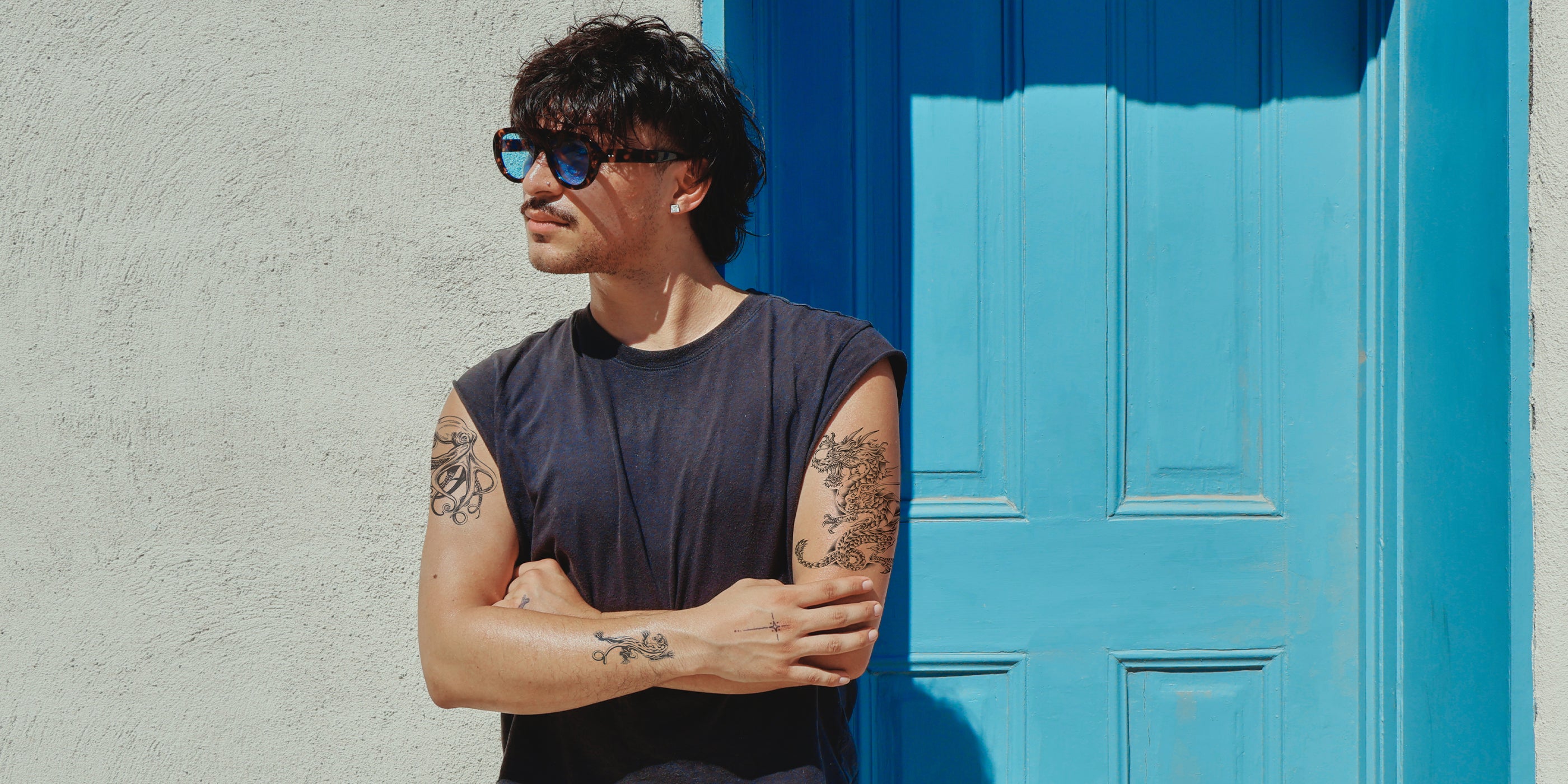 A man with dark hair and temporary tattoos wearing a sleeveless black shirt and blue-tinted sunglasses, standing with arms crossed in front of a bright blue door and textured white wall.