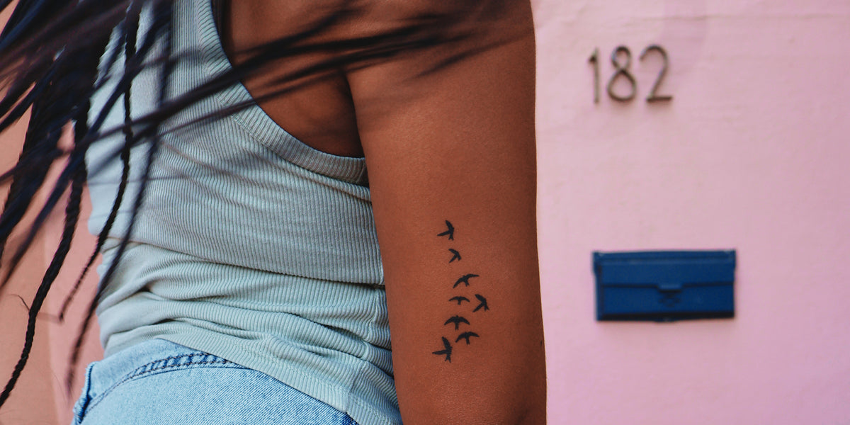A close-up of a person's upper arm featuring a small black tattoo of a flock of birds, wearing a light blue ribbed tank top and light blue jeans, standing in front of a pink wall with temporary tattoos.